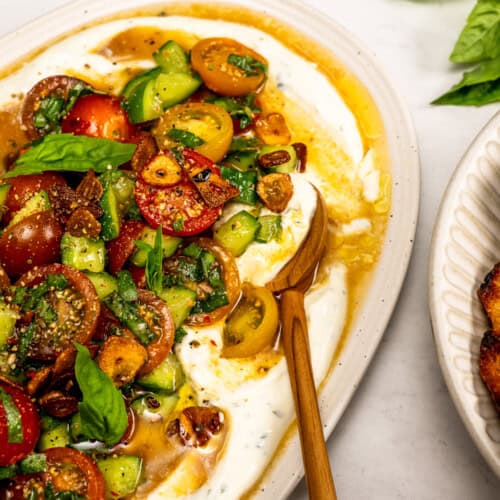 A platter with a spread of yogurt and creamy Boursin Dip, topped with a cherry tomato salad, sliced garlic, herbs, and a wooden spoon, set on a marble surface.