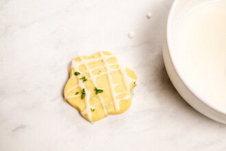 Image for recipe instruction: A Lemon Thyme Cookie with a grid pattern of white icing and decorated with thyme leaves leaves is placed next to a white bowl of icing on a light marble surface.