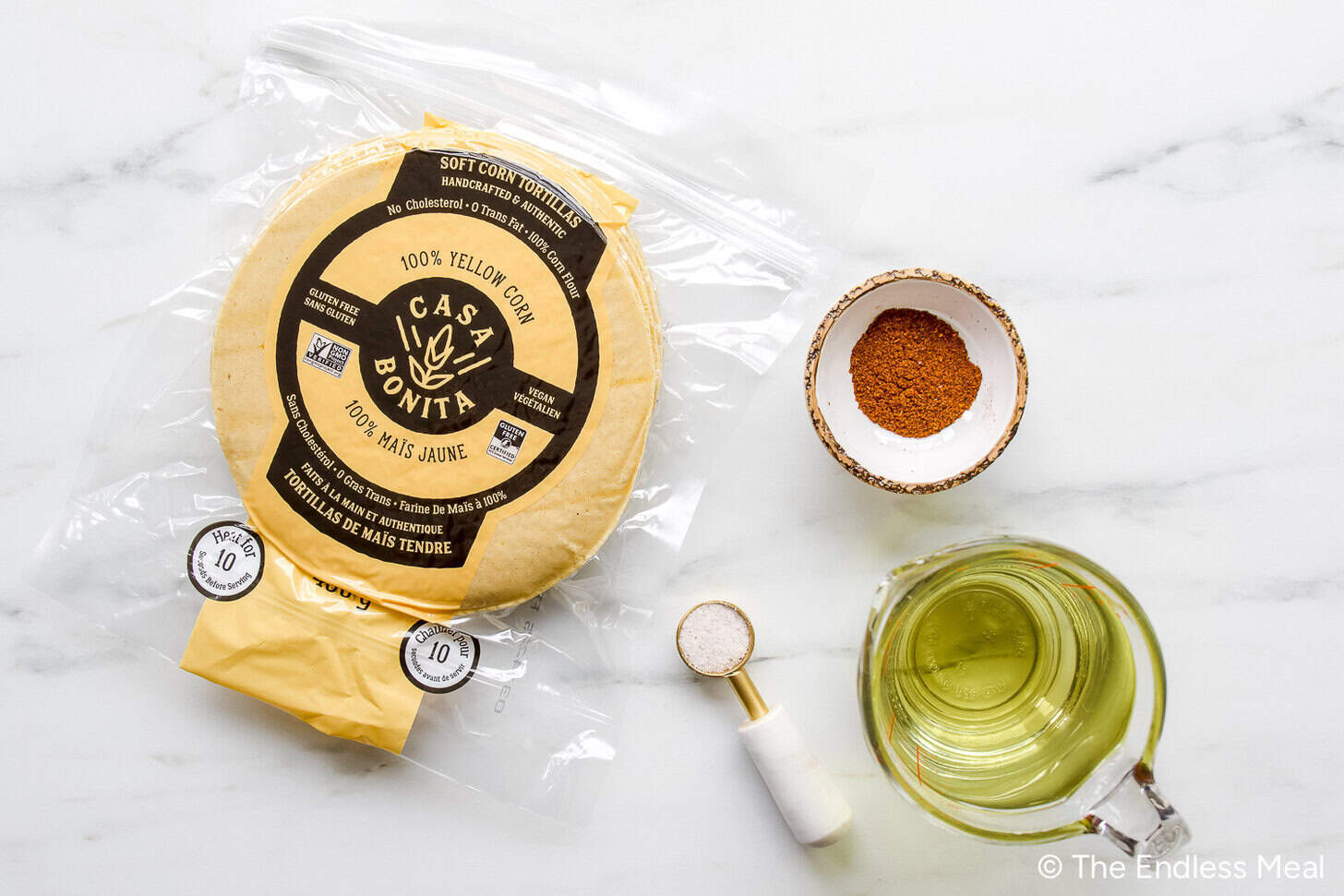 A package of yellow corn tortillas, a small bowl of chili powder, salt, and oil are ready on a white surface—perfect for making homemade tortilla chips.