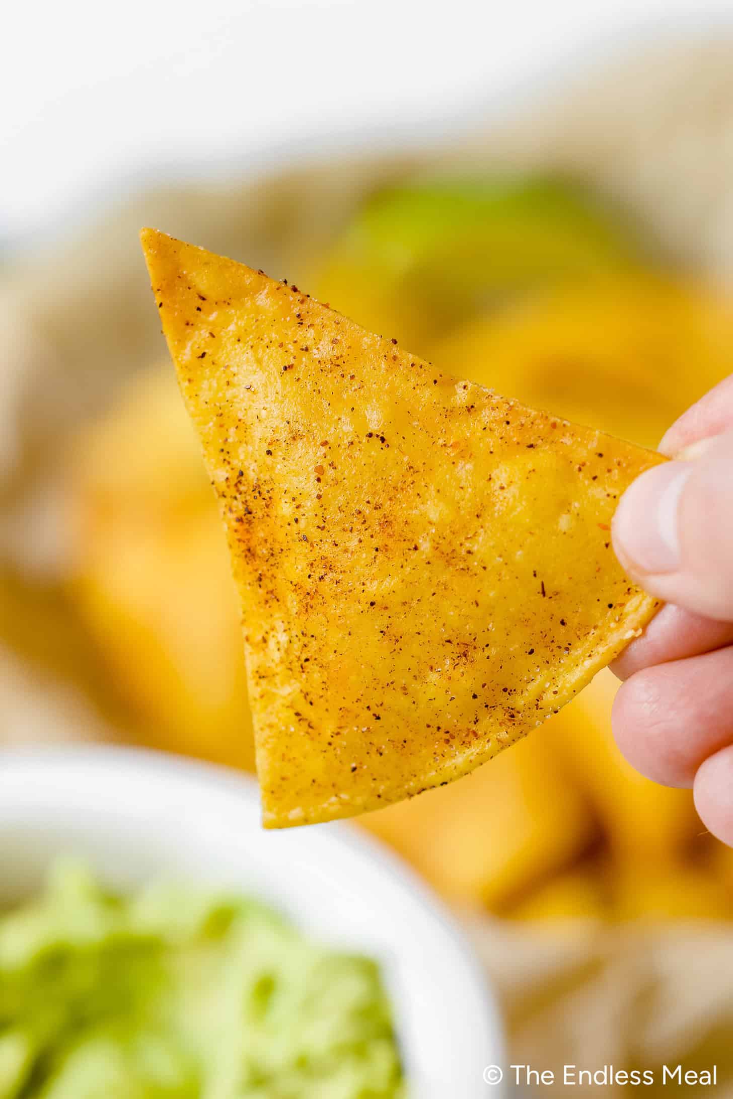 A hand holds a seasoned tortilla chip above a bowl of guacamole, with more homemade chips scattered in the blurred background.