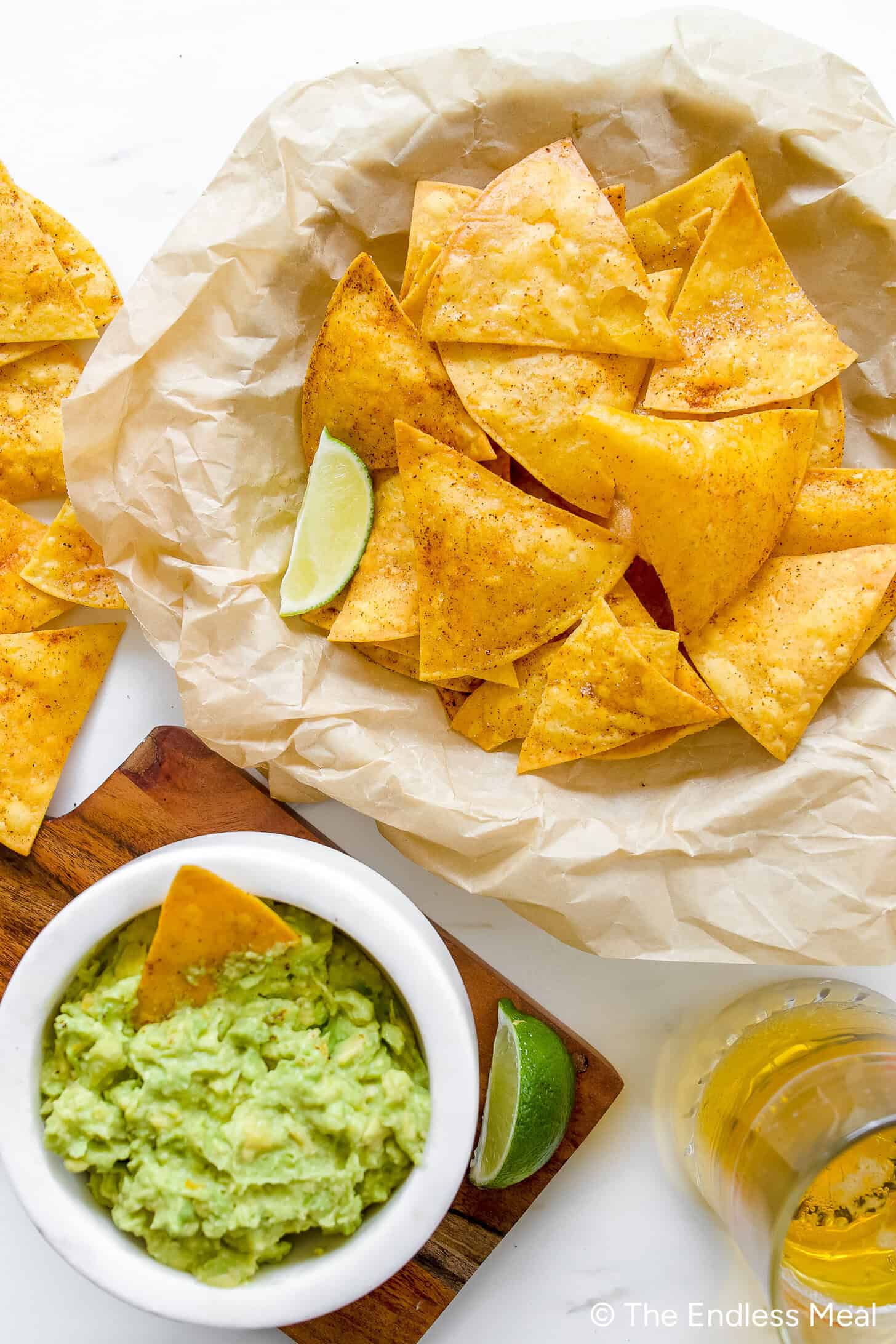A bowl of homemade guacamole with a tortilla chip, a basket of crispy tortilla chips with lime wedges on parchment paper, and a glass of beer on a white surface.