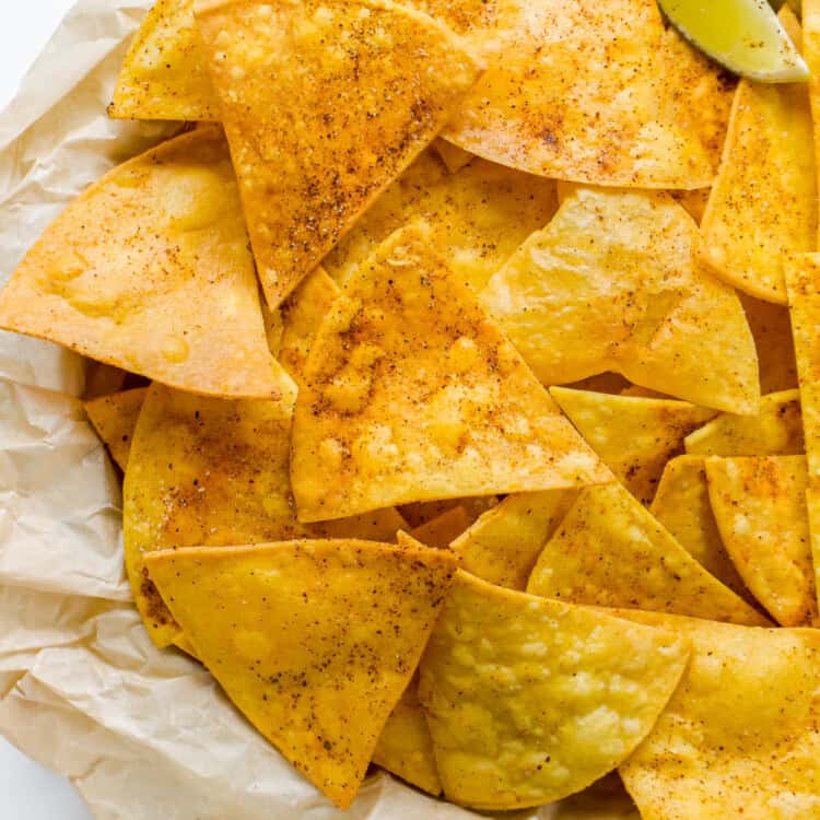 A basket of seasoned homemade tortilla chips with a lime wedge on parchment paper, served next to a small bowl of guacamole.