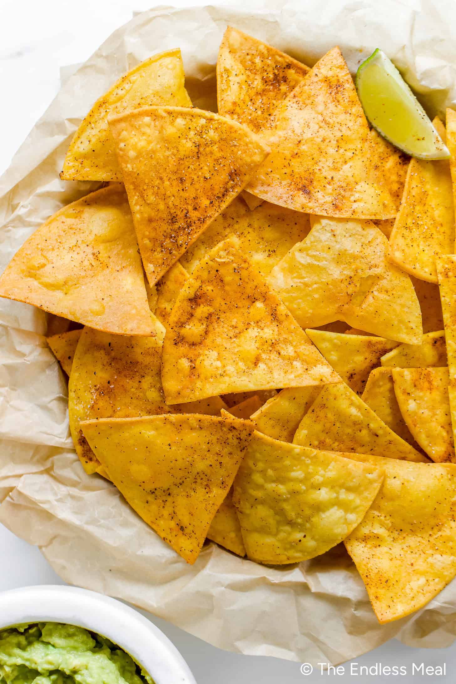A basket of seasoned homemade tortilla chips with a lime wedge on parchment paper, served next to a small bowl of guacamole.