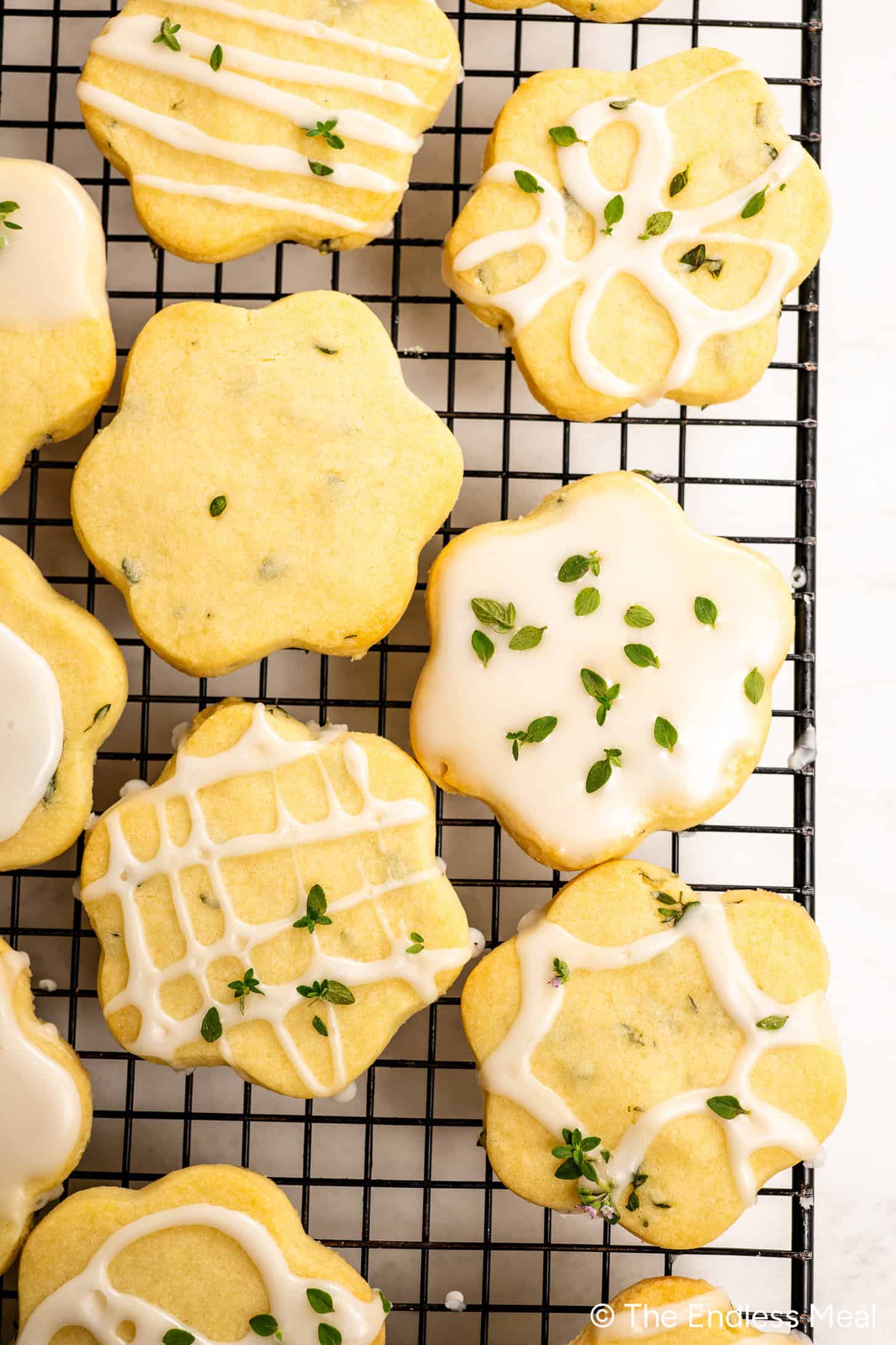 Iced Lemon Thyme Cookies with fresh thyme leaves are arranged on a black cooling rack. Some Shortbread Cookies are decorated with white icing in various patterns, highlighting the delicate blend of lemon and thyme flavors.