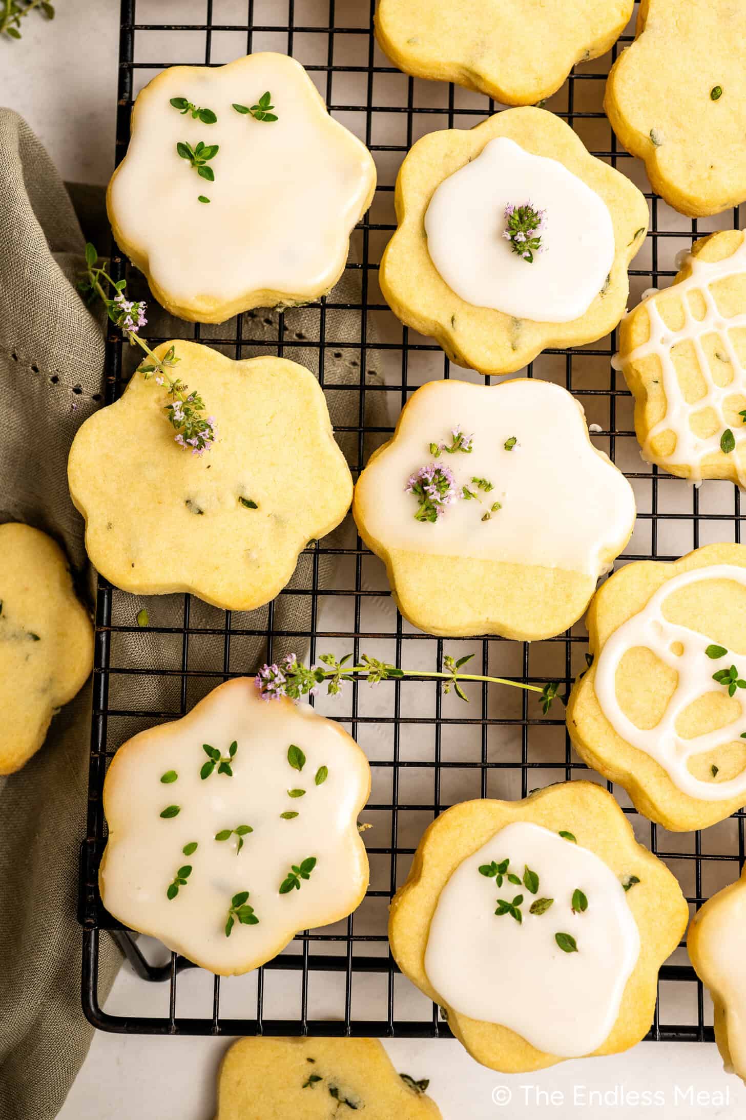 Flower-shaped Lemon Thyme Cookies with white icing and small green herb sprigs are arranged on a black cooling rack. Some shortbread cookies are plain, while others are beautifully decorated with icing and herbs.