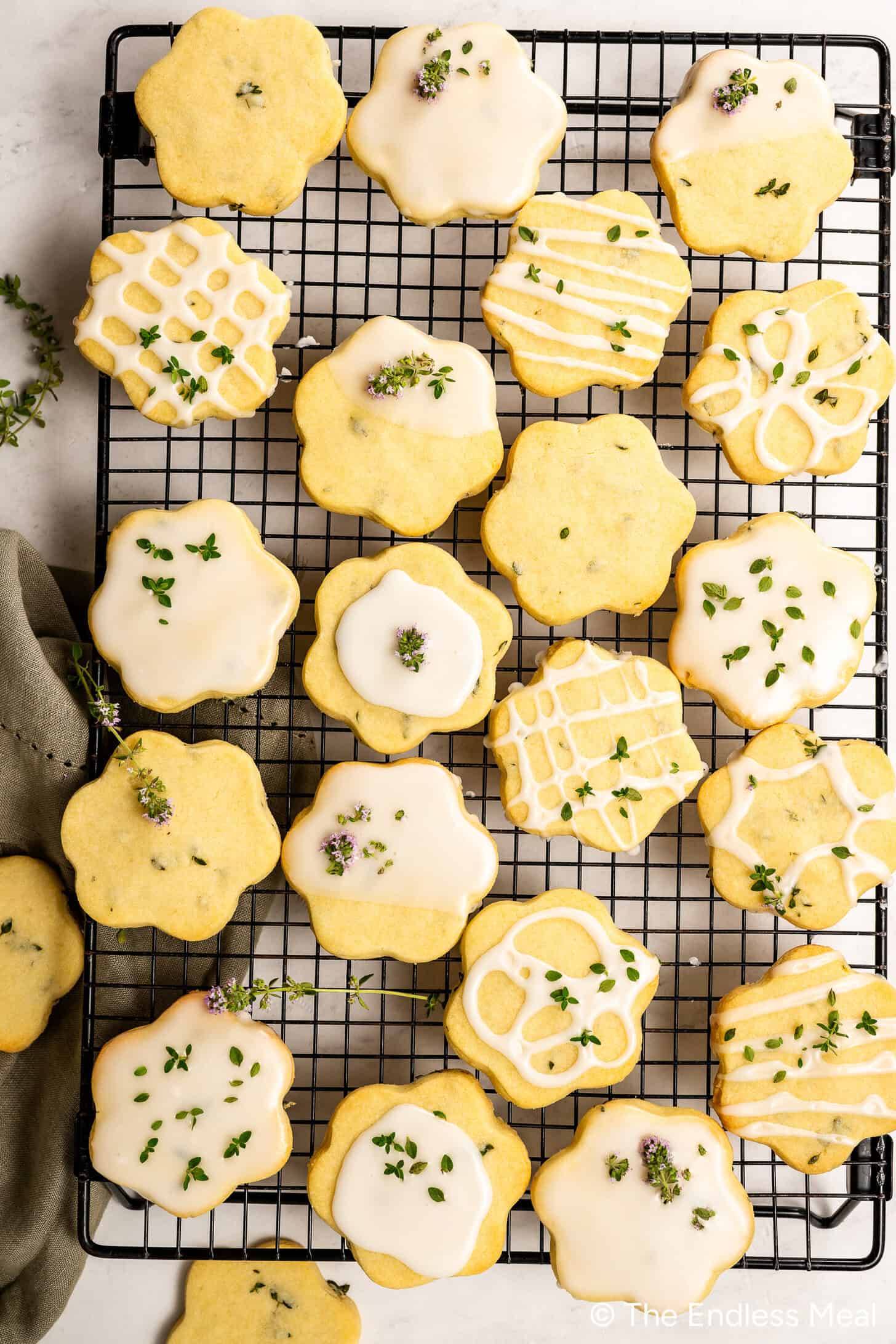 A cooling rack holds flower-shaped Lemon Thyme Cookies with white icing and fresh herb garnish; some shortbread cookies feature decorative icing patterns.