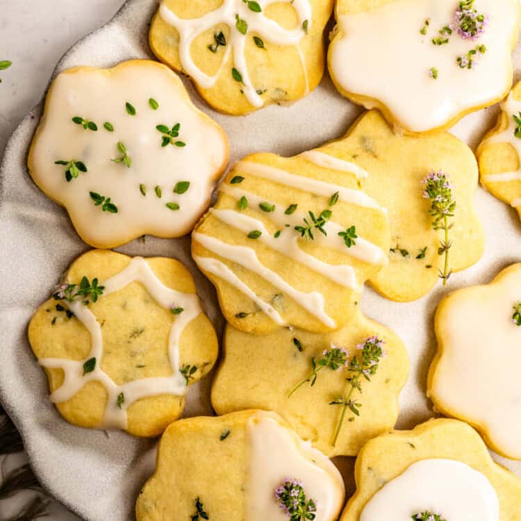 A plate of flower-shaped lemon thyme shortbread cookies with white icing, garnished with small green herbs and purple flowers.