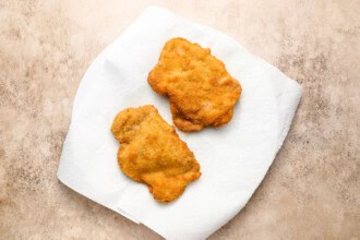Image for recipe instruction: Two breaded and fried chicken cutlets, reminiscent of a classic crispy pork cutlet, rest on a paper towel atop a white plate on a beige surface.