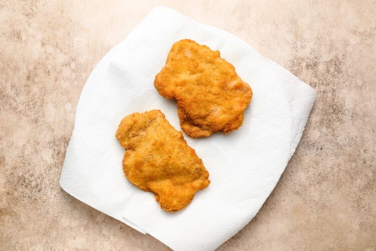 Image for recipe instruction: Two breaded and fried chicken cutlets, reminiscent of a classic crispy pork cutlet, rest on a paper towel atop a white plate on a beige surface.