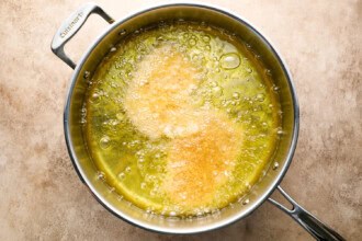Image for recipe instruction: A stainless steel pan with hot oil frying two crispy pork cutlets, creating bubbles and golden brown spots, on a beige countertop.