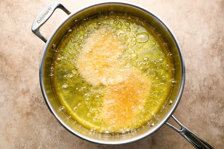 Image for recipe instruction: A stainless steel pan with hot oil frying two crispy pork cutlets, creating bubbles and golden brown spots, on a beige countertop.