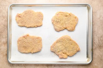 Image for recipe instruction: Four crispy pork cutlets are arranged on a parchment-lined baking sheet, ready to be cooked.