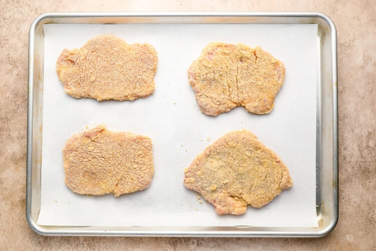 Image for recipe instruction: Four crispy pork cutlets are arranged on a parchment-lined baking sheet, ready to be cooked.