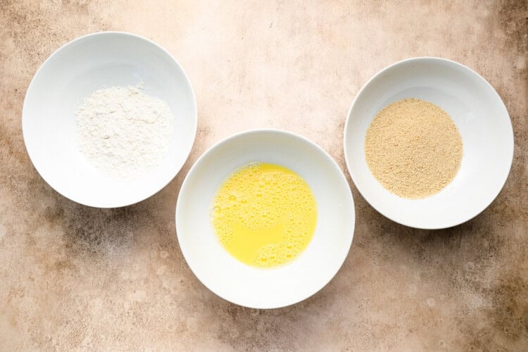 Image for recipe instruction: Three white bowls on a beige surface contain flour, beaten egg, and breadcrumbs arranged in a triangle—essential for preparing a delicious crispy pork cutlet.