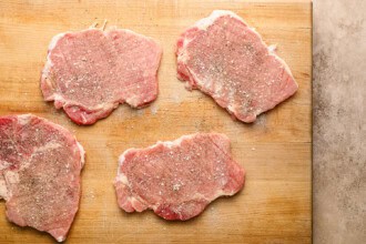 Image for recipe instruction: Four raw pork chops sprinkled with salt and pepper are arranged on a wooden cutting board, ready to be transformed into a delicious crispy pork cutlet.