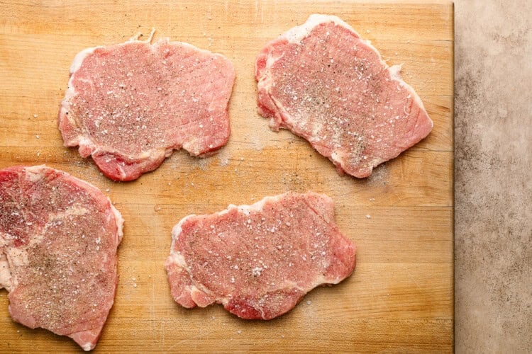 Image for recipe instruction: Four raw pork chops sprinkled with salt and pepper are arranged on a wooden cutting board, ready to be transformed into a delicious crispy pork cutlet.
