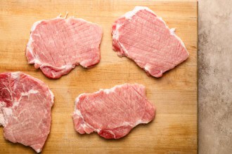 Image for recipe instruction: Four raw pork chops on a wooden cutting board, ready to be transformed into a delicious crispy pork cutlet.