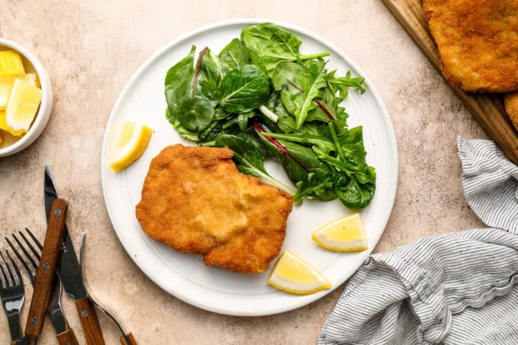 Image for recipe instruction: A plate with a crispy pork cutlet, a small green salad, and lemon wedges. Utensils, a napkin, and a bowl of lemon are nearby.