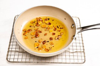 Image for recipe instruction: A frying pan with sliced garlic browned in oil, perfect for topping Boursin dip with marinated tomatoes, rests on a metal cooling rack on a white surface.