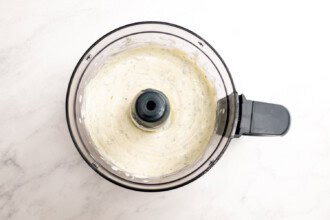 Image for recipe instruction: A food processor bowl containing creamy Whipped Boursin sauce with visible green herb flecks sits on a white surface.