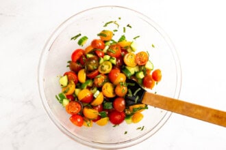 Image for recipe instruction: A clear glass bowl filled with a salad of marinated tomatoes, chopped cucumber, and herbs, with a wooden spoon resting inside ready for a topping for Boursin dip.
