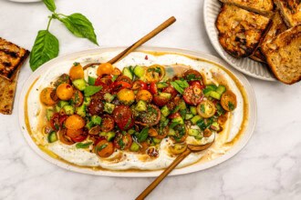 Image for recipe instruction: An oval platter with Whipped Boursin cheese dip topped with marinated tomatoes, cucumbers, herbs, and spices, served with grilled bread on the side.