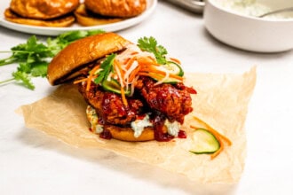 A Gochujang Fried Chicken Sandwich with cilantro mayo, pickled vegetables, and cucumbers on a toasted bun, served on parchment paper.