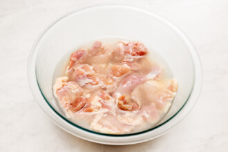 Raw chicken pieces soaking in a glass bowl filled with water on a white countertop, ready to be transformed into a delicious Gochujang Fried Chicken Sandwich.