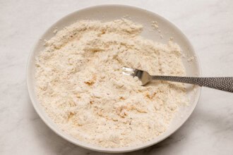 A white bowl containing flour mixture with a silver fork resting inside, ready for making a crispy Fried Chicken Sandwich, is placed on a light-colored surface.