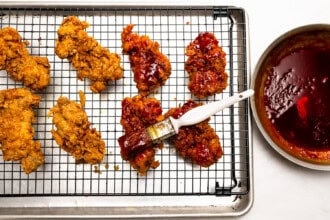 Gochujang Fried Chicken pieces on a wire rack, some being brushed with red gochujang sauce, next to a bowl filled with more sauce—perfect for stacking in a sandwich or dipping in cilantro mayo.