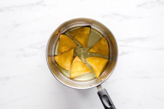 Image for recipe instruction: Six homemade tortilla chips are frying in hot oil inside a silver saucepan with a black handle, placed on a white marble surface.