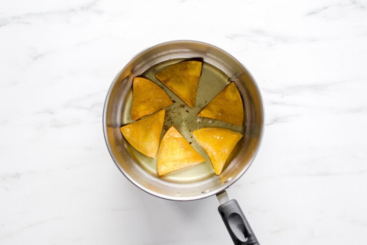 Image for recipe instruction: Six homemade tortilla chips are frying in hot oil inside a silver saucepan with a black handle, placed on a white marble surface.