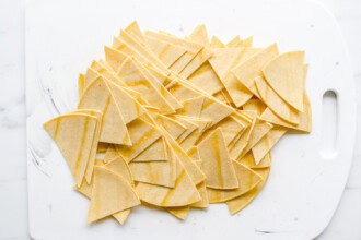 Image for recipe instruction: Corn tortillas cut into triangles are spread out on a white cutting board with a handle, ready to be baked or fried into crispy homemade tortilla chips.