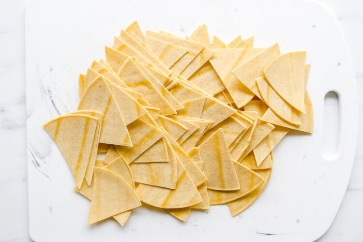 Image for recipe instruction: Corn tortillas cut into triangles are spread out on a white cutting board with a handle, ready to be baked or fried into crispy homemade tortilla chips.