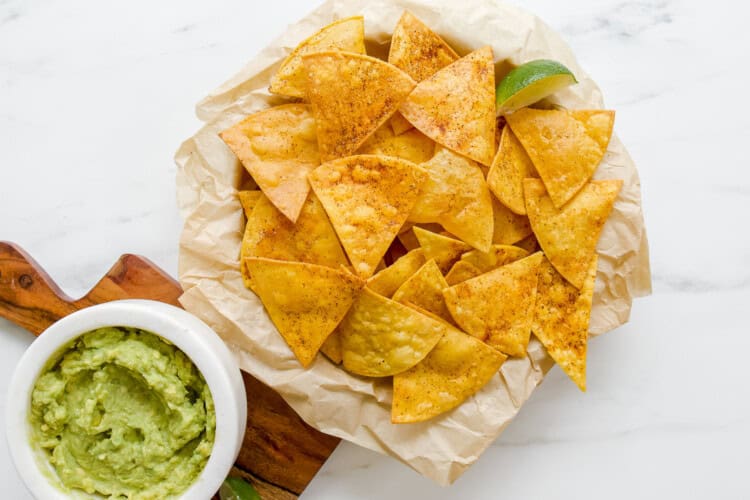 Image for recipe instruction: A plate of seasoned homemade tortilla chips on parchment paper with a wedge of lime, next to a bowl of guacamole on a wooden board.