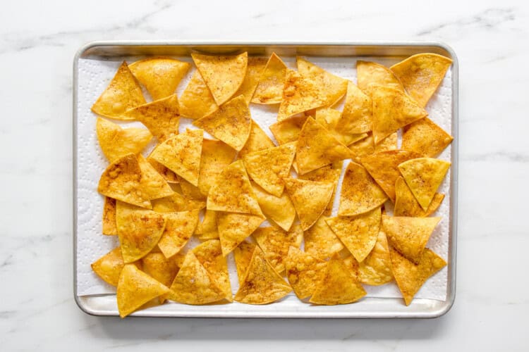 Image for recipe instruction: A baking sheet lined with paper towels holds evenly spread, golden-brown homemade tortilla chips.