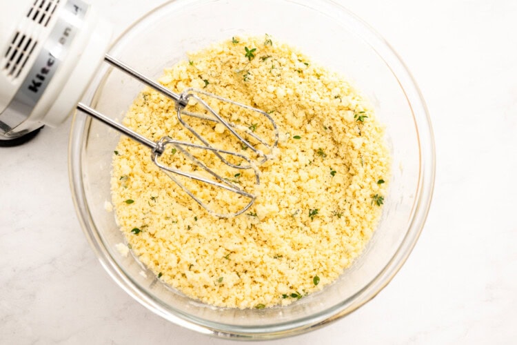 Image for recipe instruction: A glass bowl contains crumbly yellow Lemon Thyme shortbread dough with green herbs being mixed by an electric hand mixer on a white surface.