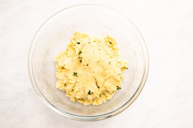 Image for recipe instruction: A clear glass bowl containing a mound of pale yellow dough with flecks of thyme, perfect for making lemon thyme shortbread cookies, placed on a white surface.