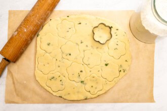 Image for recipe instruction: Rolled-out dough for Lemon Thyme Cookies with chopped herbs is being cut into flower shapes using a cookie cutter; a rolling pin and jar of flour rest nearby on parchment paper.