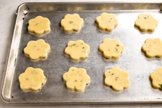 Image for recipe instruction: A metal baking sheet with twelve uncooked, flower-shaped Lemon Thyme Shortbread cookie dough pieces spaced evenly apart, ready to be baked.