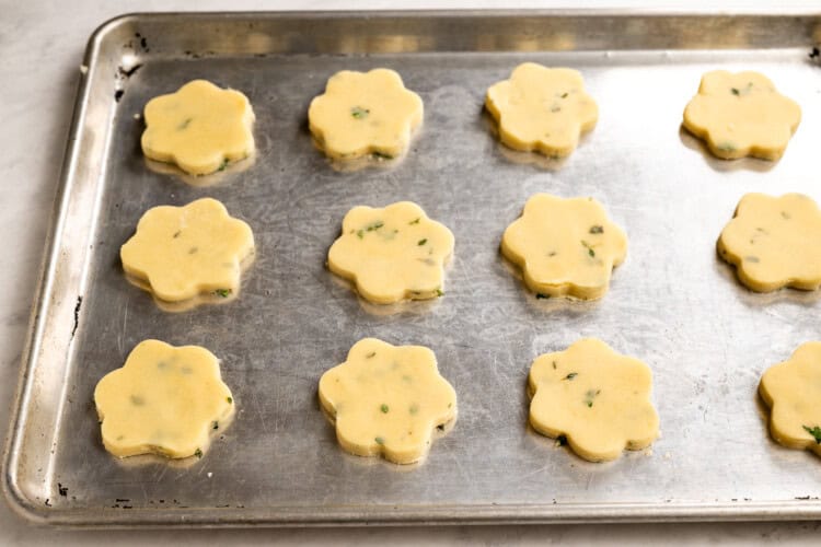 Image for recipe instruction: A metal baking sheet with twelve uncooked, flower-shaped Lemon Thyme Shortbread cookie dough pieces spaced evenly apart, ready to be baked.