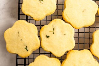 Image for recipe instruction: Six flower-shaped Lemon Thyme Cookies with green herb pieces are cooling on a black wire rack set on a light-colored surface.
