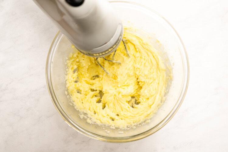 Image for recipe instruction: A stand mixer is beating a yellow mixture for Lemon Thyme Cookies in a glass bowl on a white countertop.