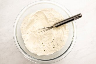 Image for recipe instruction: A metal whisk rests in a glass bowl filled with flour and a few thyme leaves, ready for making Lemon Thyme Shortbread, on a white surface.