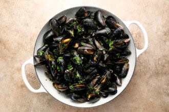 Image for recipe instruction: A white pan filled with cooked mussels garnished with chopped parsley, placed on a light textured surface—perfect for pairing with crispy frites for a classic moules frites experience.