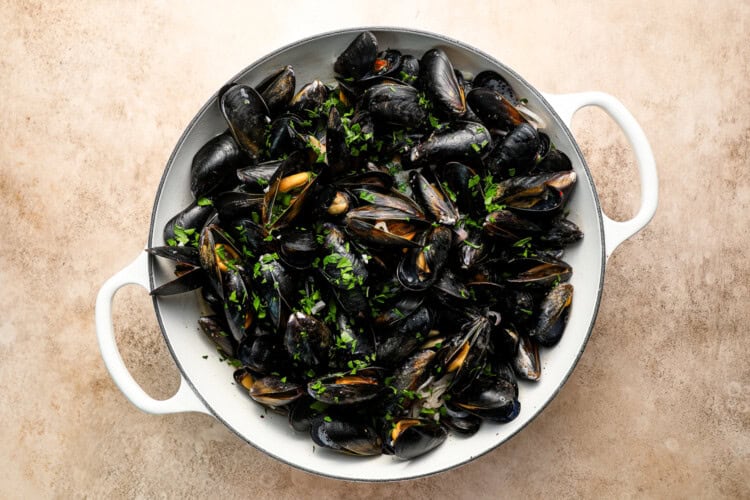 Image for recipe instruction: A white pan filled with cooked mussels garnished with chopped parsley, placed on a light textured surface—perfect for pairing with crispy frites for a classic moules frites experience.