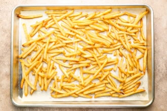 Image for recipe instruction: A baking sheet lined with parchment paper holds a single layer of baked golden French fries, perfect for serving alongside classic Moules Frites.