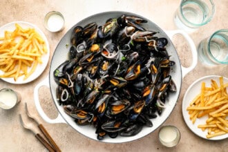 Large white pan filled with cooked mussels sits at the center, surrounded by plates of crispy French fries—classic Moules Frites—plus glasses of water and dipping sauces on a light table.