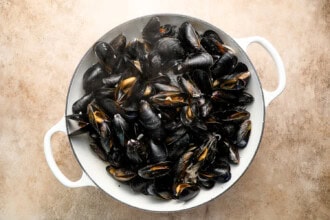 Image for recipe instruction: A white pot filled with cooked black mussels, some shells open, shown from above on a light-colored surface—a classic Moules Frites presentation.