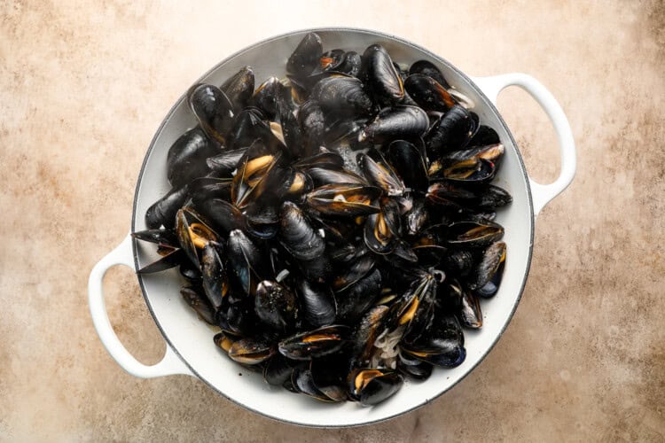 Image for recipe instruction: A white pot filled with cooked black mussels, some shells open, shown from above on a light-colored surface—a classic Moules Frites presentation.
